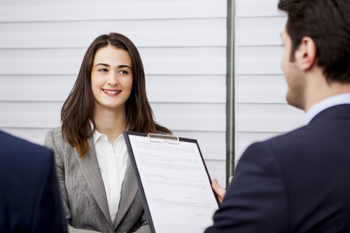 Eine Frau wird im Vorstellungsgespräch von zwei Männern interviewt, einer hält die Bewerbungsunterlagen in den Händen